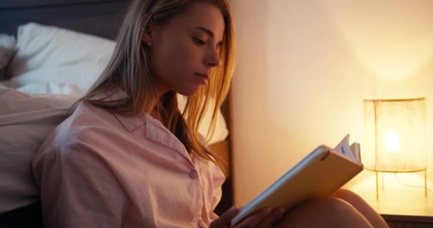 ピンクのナイトガウンのブロンドの女の子がランプのついた本を読んでいます 読書前にベッドに行く — ストック動画