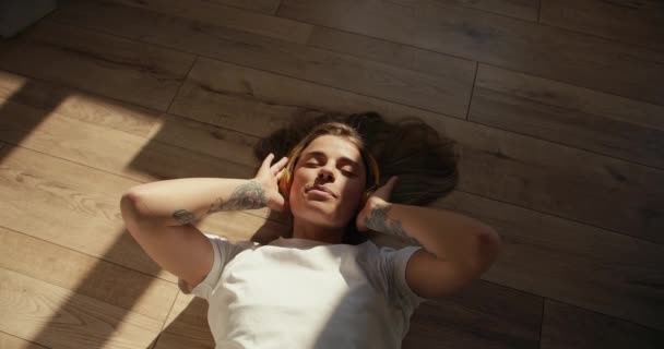 闭着眼睛的金发女孩躺在地板上 用无线耳机听音乐 — 图库视频影像