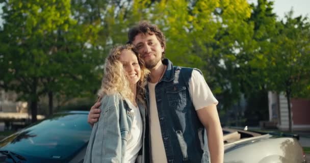 美しい若いカップルブロンドの女の子とブルネットの男の肖像画は灰色の車の背景にポーズをとっています 素敵な車で幸せなカップル — ストック動画