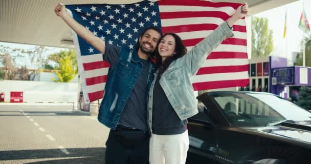 一对穿着皮夹克的年轻夫妇和一个黑发女孩笑着看着摄像机 一个男的和一个女的把美国国旗举过头顶 站在一辆灰色敞篷车旁边 旁边是一辆汽油 — 图库视频影像