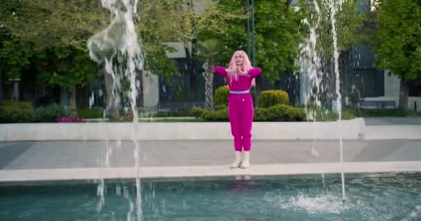一个穿着粉色衣服 一头粉色头发的女孩在靠近喷泉的街上做瑜伽 快乐积极地休息在户外聪明的女孩 — 图库视频影像