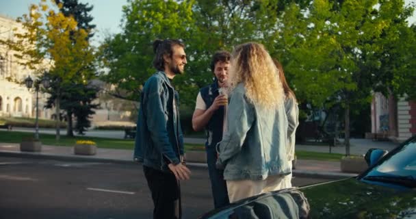 在阳光灿烂的一天 一群穿着牛仔夹克的年轻朋友站在他们灰色的出租车旁边 喝着黄色的鸡尾酒 — 图库视频影像