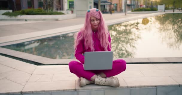 一个穿着粉色衣服 一头粉色头发的女孩在喷泉边的笔记本电脑上工作 室外远程工作 — 图库视频影像