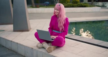 Pembe elbiseli ve pembe saçlı mutlu bir kız sokakta bir çeşmenin yanında otururken laptopun başında neşeli bir şekilde çalışıyor..