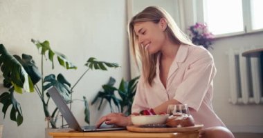 Açık pembe elbiseli sarışın bir kız laptopta çalışıyor ve güneşli bir odada kahve içiyor..