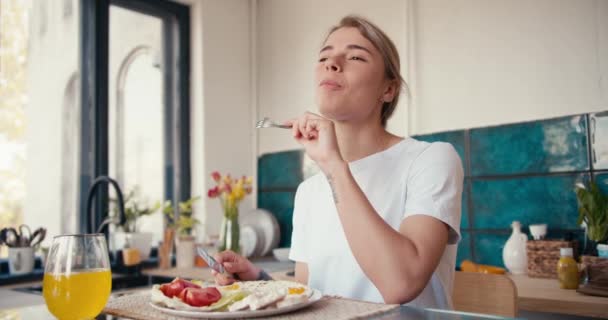 白いTシャツを着たブロンドの女の子が 晴れた朝にキッチンで彼女の国内朝食を楽しんでいます 適切な栄養は幸せな生活の基盤です — ストック動画