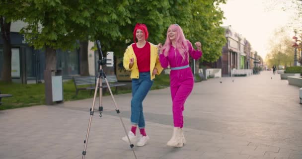两个穿着五颜六色衣服的快乐姑娘在街上跳着同步舞 女孩们在三脚架上的手机上拍照 — 图库视频影像
