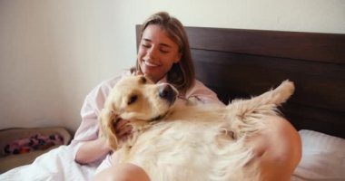 Beyaz gecelikli sarışın bir kız köpeğine sarılıyor, açık renkli, yatakta yatıyor..