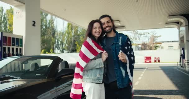 革のジャケットを着た若い男とブルネットの女の子のカップルが笑っていて カメラを見て 彼らは米国の旗に包まれています — ストック動画