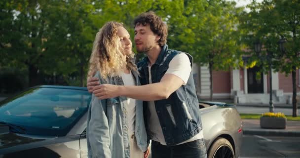 在阳光明媚的夏日 一对年轻貌美的黑发男人和一个金发姑娘站在那里 看着摄像机 靠着灰色出租车的车尾拥抱在怀里 — 图库视频影像