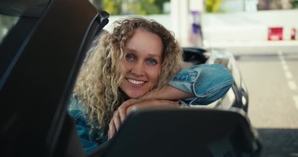 站在加油站一辆深灰色出租车的前座上 美丽的金发姑娘 蓝眼睛 摆出姿势 看着镜头 — 图库视频影像