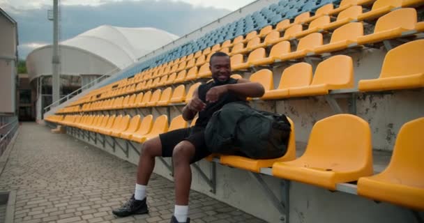 黒のスポーツの夏の制服の黒い肌の選手は 彼のバッグから水の特別なボトルを取り出し スタジアムのスタンドでそれを飲む — ストック動画