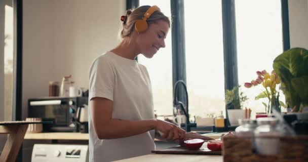 黄色のヘッドフォンと白いTシャツのブロンドの女の子は音楽を聞き 彼女の朝食を準備します — ストック動画