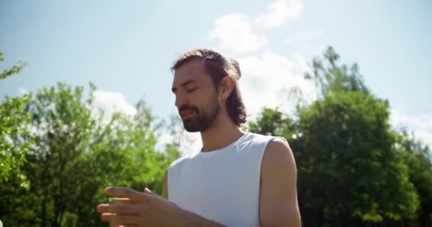 緑の木々と青い空を背景に自然の中で水を飲む白いTシャツのブルネットの男の概要 屋外スポーツをプレイした後の休息と休憩 — ストック動画