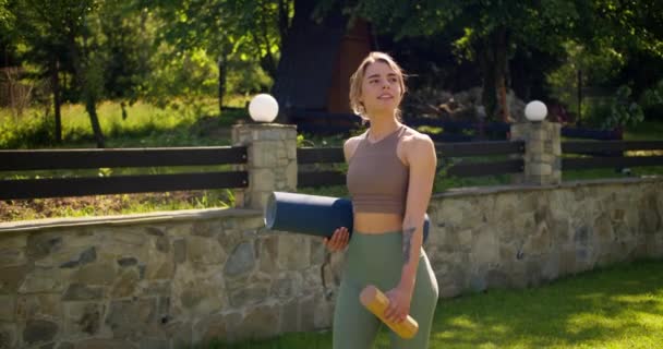 一个身穿夏季运动服的金发女孩沿着石墙边的绿色草坪走着 手里拿着卡里玛和一瓶水 体育运动和瑜珈 — 图库视频影像