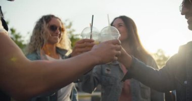 Bir grup modaya uygun kot ceketli genç, güneşli bir akşamda sokakta sarı bir kokteyl ile plastik bardaklarını tokuşturuyor..