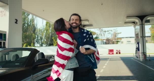ハッピーブルネット女の子接吻彼女のボーイフレンド カップルラップで米国フラグとスタンドで彼らのグレーコンバーチブル近くのガソリンスタンド — ストック動画