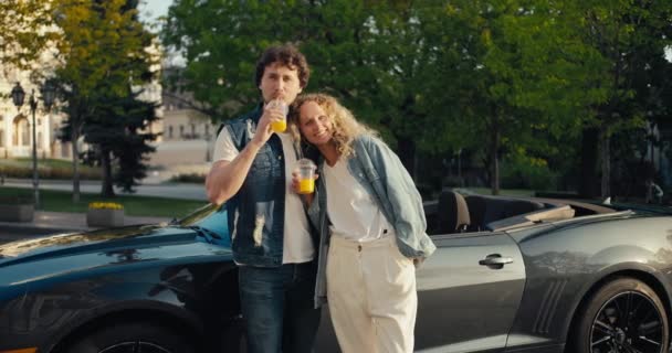 一个男的和一个女的站在一辆灰色的出租车旁边笑着 看着摄像机 喝着黄色的鸡尾酒 — 图库视频影像
