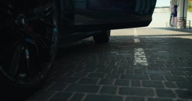 Beyaz spor ayakkabılı bir adamın üstü açık koyu gri bir arabadan inip benzin istasyonunda arabasına benzin doldurduğu yakın plan bir fotoğraf..