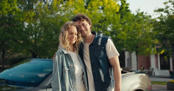 若いですブルネットの男とブロンドの女の子の肖像画で彼らの灰色の車の近くに立っているデニムジャケット笑顔と夏の美しい木を背景にカメラを見て — ストック動画