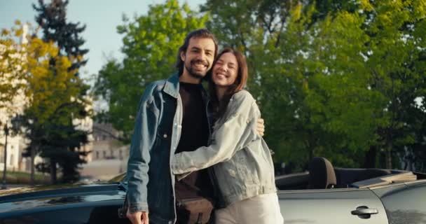 幸せなカップルの男とブルネットの女の子でデニムジャケットスタンド 抱擁と灰色のCabrioletと木の背景にカメラを見て — ストック動画