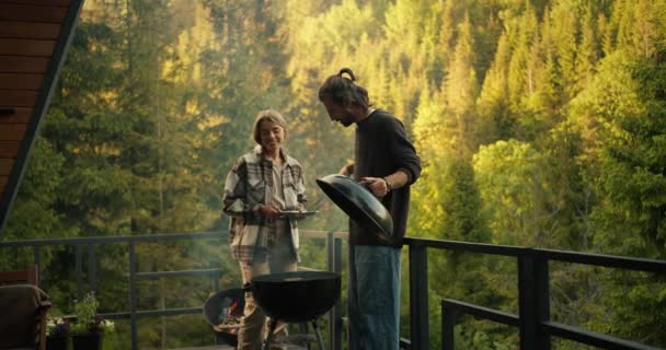 緑豊かな針葉樹林を背景に 若いカップル 男と女の子は田舎の家でリラックスして特別なグリルで肉を揚げています — ストック動画