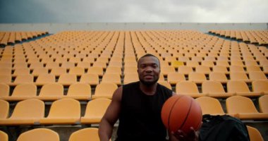 Siyah formalı genç bir atlet stadyumun sarı tribünlerinde oturuyor kameraya bakıyor ve elinde bir basketbol topu tutuyor..