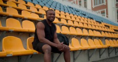 Siyah spor üniformalı bir atlet stadyumun tribünlerinde oturur, kameraya bakar ve gülümser..