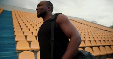 Siyah tişörtlü, siyah tenli bir sporcunun, tribünlerde sarı sandalyelerle stadyumda yürüyen bir çantasının görüntüsü..