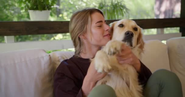 一个穿着深红色毛衣的金发姑娘坐在沙发上 抱着她那只浅色的狗抚摸着 — 图库视频影像