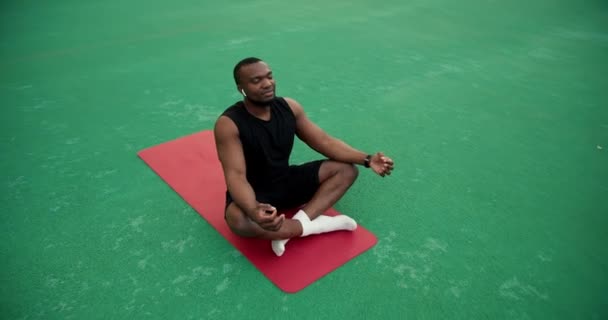 黒いスポーツの夏の服の黒い肌の色の男は赤いマットの上に座ってスタジアムで瞑想 — ストック動画