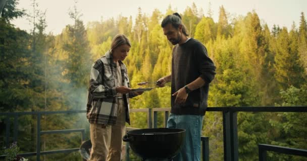 茶色のセーターのブルネット男とブロンドの女の子フライバーベキュー肉 統一された森のカントリーハウスのバルコニー上の特別なグリルで — ストック動画