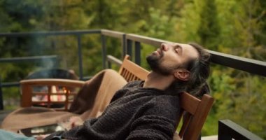 A young brunette man with closed eyes is resting on a sofa on the balcony of a country house overlooking the mountains and a coniferous forest, firewood is burning near him in the barbecue.