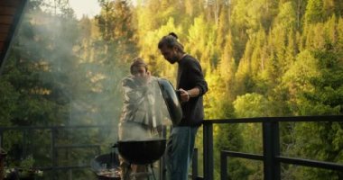 A brunette guy and a blonde girl are watching the preparation of meat on the grill during a picnic on the terrace against the backdrop of a coniferous forest and mountains.