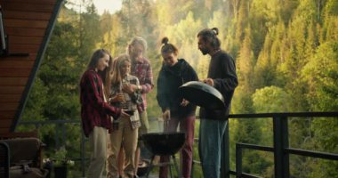 Friends at a picnic: a girl puts sausages on a wood-fired grill, her friends help her in this. Barbecue on the balcony of a country house in the mountains with a coniferous forest.