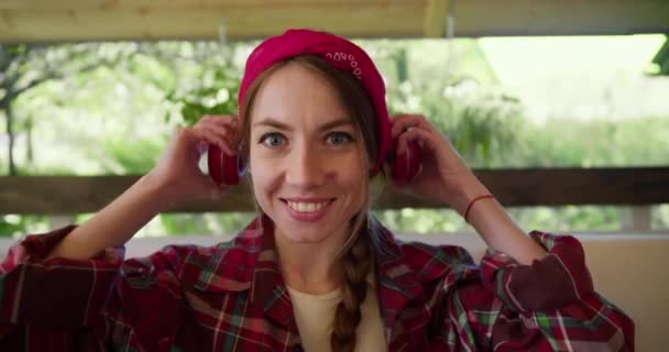 一个穿着红色格子花衬衫的女孩 穿着红色的无线耳机 听音乐 摇着头坐在沙发上 就像在看电影一样 — 图库视频影像