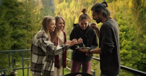 一群朋友在一个可以俯瞰高山的乡间小木屋里 用针叶林在烤架上做的食物 那家伙拿着一盘食物站在烤肉架旁边 — 图库视频影像