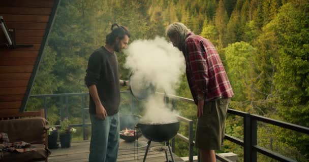 两个人在烤架上检查他们做的食物 互相吹5下 因为他们做得很好 浓重的白烟从火盆里冒出 映衬着高山和大海 — 图库视频影像