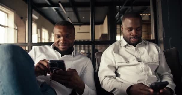 白いシャツの黒い肌を持つ2人の従業員は アームチェアのオフィスに座ってスマートフォンで作業します — ストック動画