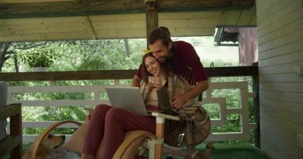 一个穿着红色T恤的黑发男人围住一个坐在扶手椅上的女孩 看着笔记本电脑屏幕 在森林的背景下喝茶 对儿童的照料的表现 — 图库视频影像