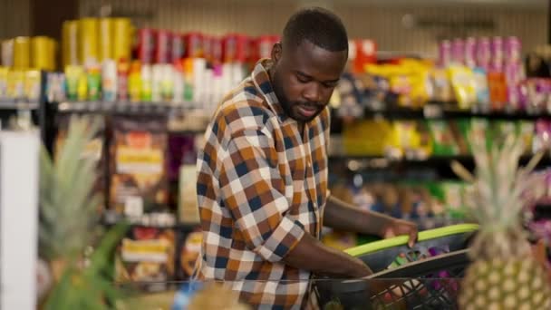 スーパーマーケットのフルーツカウンターに沿って歩き 正しい製品を選択する平凡なシャツの黒い肌の色を持つ男 — ストック動画