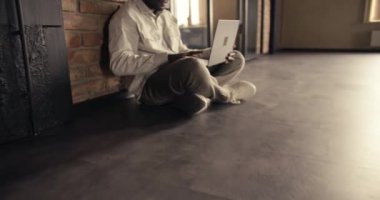 The camera zooms in and takes a look at an office worker who sits on the floor in a white shirt and works behind a laptop in a loft-style office against a brick wall.