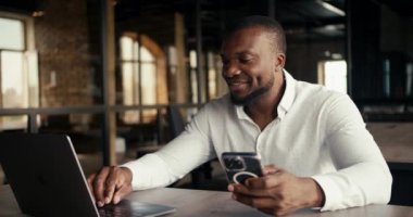 A man with a Black skin color works in the office at the same time on a laptop and on a phone.