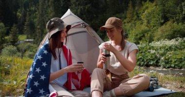 Şapkalı sarışın bir kız, Amerika Birleşik Devletleri bayrağına sarılmış esmer bir kız için yeşil bir ormanın arka planına karşı çay koyuyor..