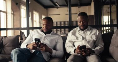 2 employees with Black skin office workers in white shirts sit on the couch communicate and chat on smartphones.