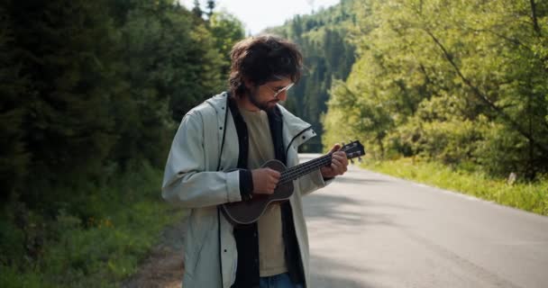 道路の近くの白いジャケットのブルネット男は 山のハイキングで音楽弦楽器を演奏します — ストック動画