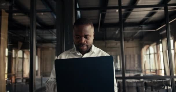 一个穿着白衬衫 手里拿着笔记本电脑的黑皮肤男人 在办公室里走来走去 穿着阁楼风格的衣服 — 图库视频影像