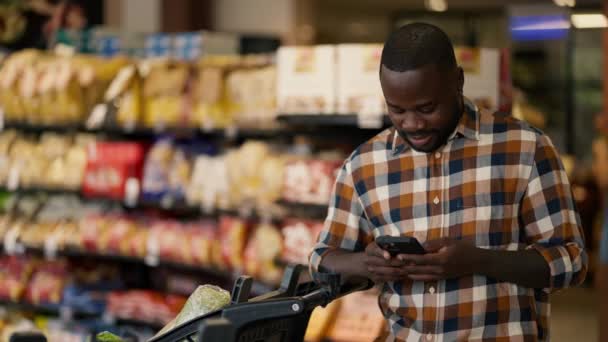 一个黑皮肤的男人穿着格子呢衬衫 一边用手机打字一边聊天 旁边是超级市场的一辆购物车 — 图库视频影像