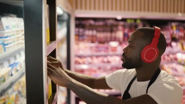 特写镜头 一个黑皮男子头戴红色耳机 一条黑色围裙在超级市场的货架上调整商品 — 图库视频影像