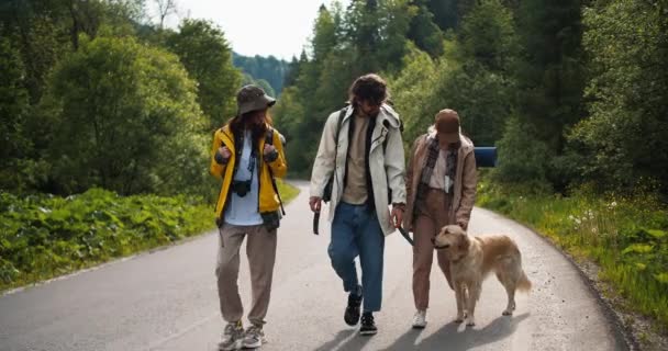山岳地帯の森沿いの道を歩いている彼らの光色の犬と一緒にハイキングのための特別な服の男と2人の女の子 — ストック動画